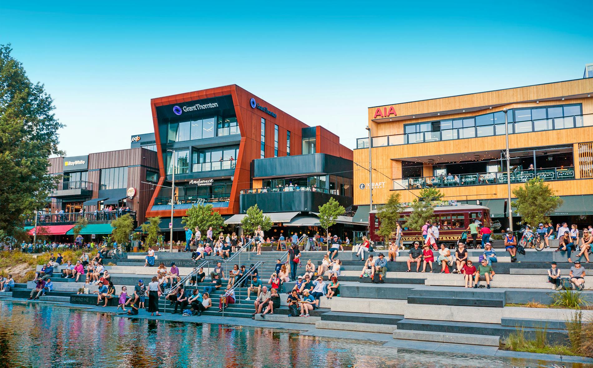 Te Pae hosts a vibrant scene where people relax on steps by a waterway, enjoying the surrounding modern buildings and green spaces, creating a lively atmosphere for social engagement.